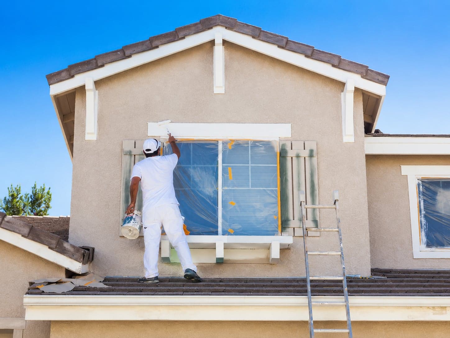 A professional painter in white work clothes, seen from behind, rolling paint onto the exterior window trim of a stucco house