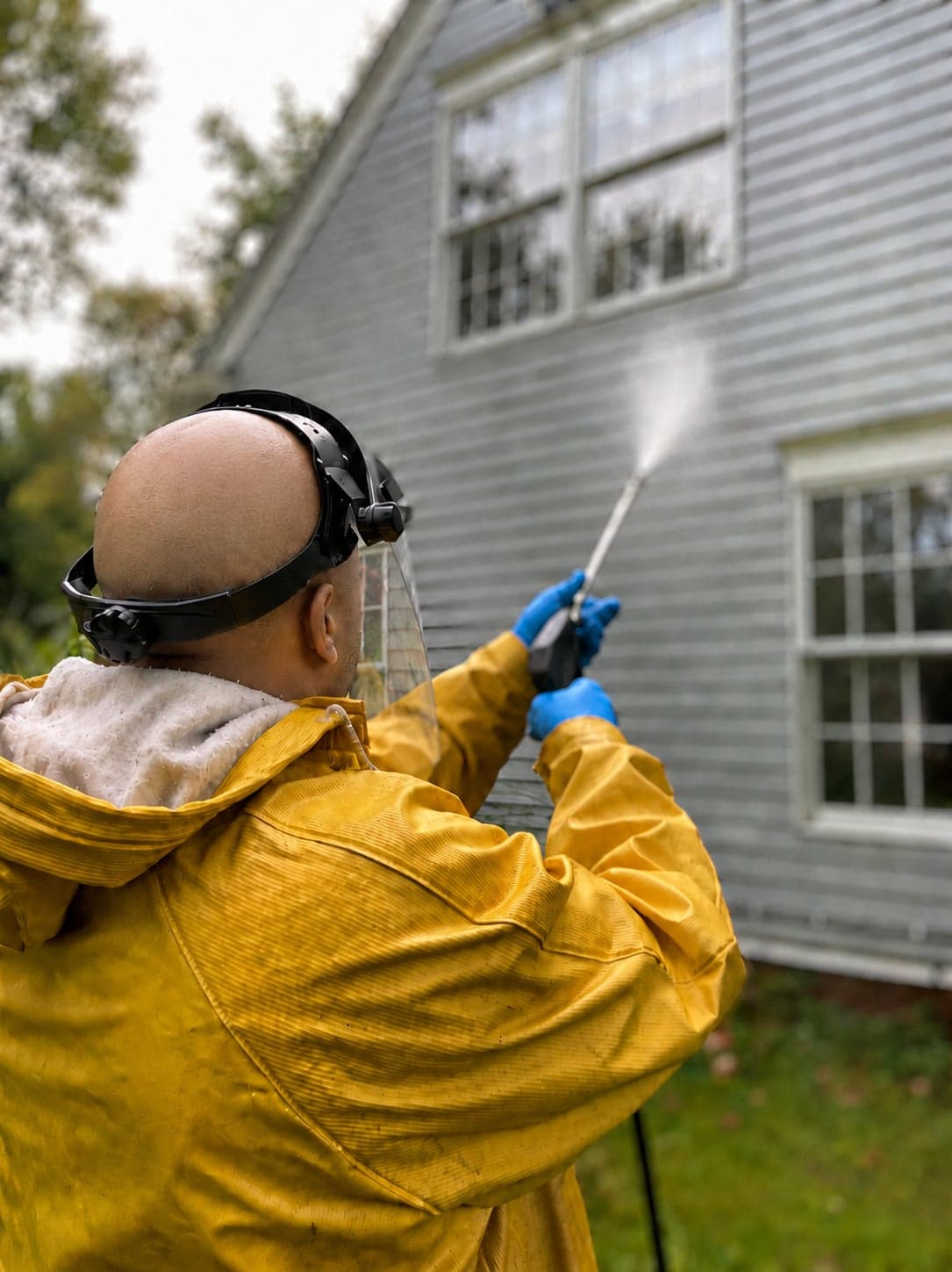 A worker in a yellow rain jacket, face shield, and gloves power washing the clapboard siding of a house